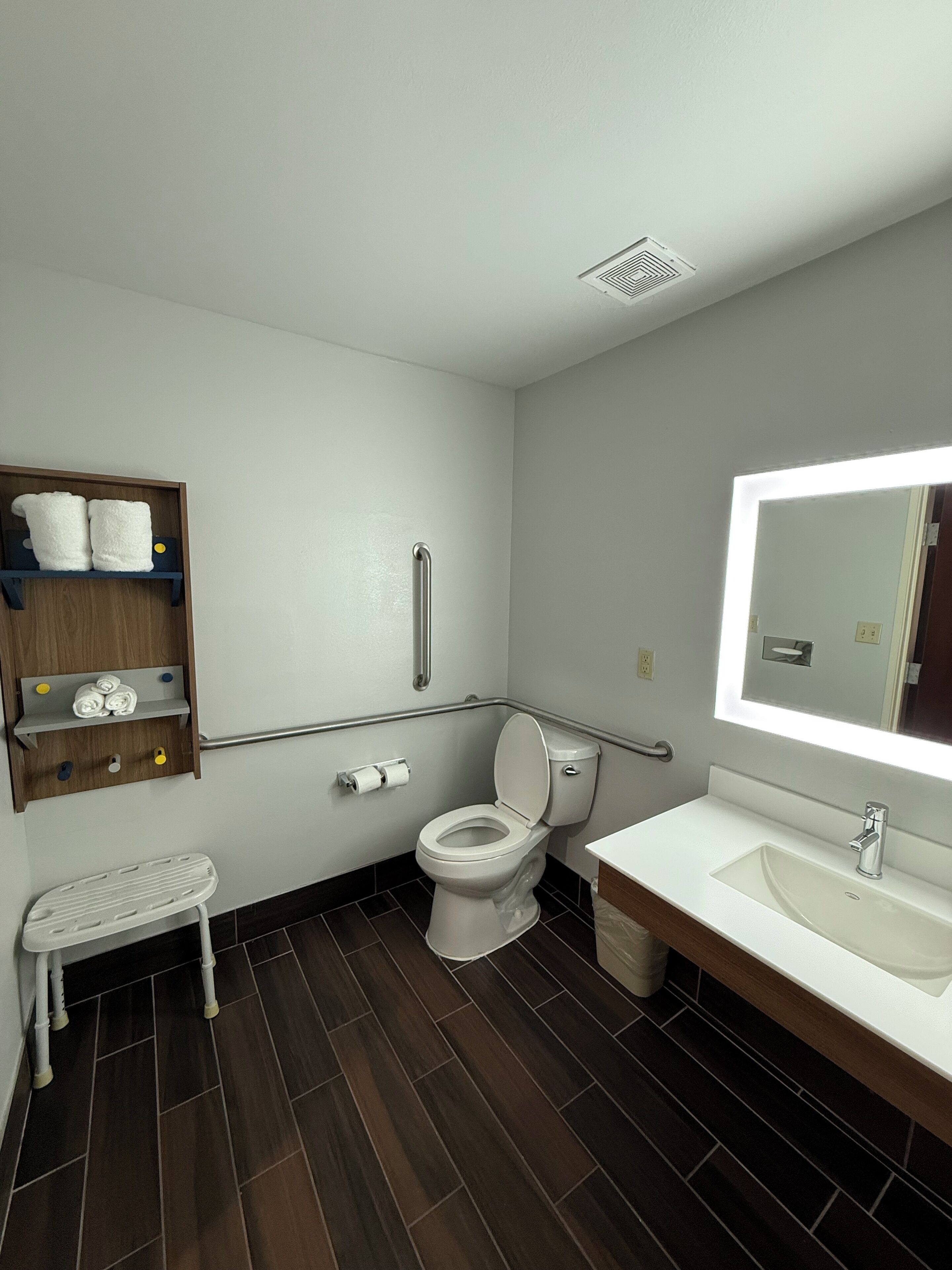 Ein modernes Badezimmer mit einer weißen Toilette, einem Waschbecken und einem Spiegel. Eine Ablagefläche mit Handtüchern und ein kleiner Tritthocker sind vorhanden.