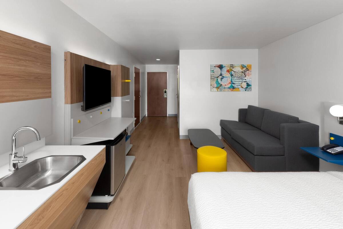 A modern hotel room with a kitchenette, a gray sofa, a bed, and a small yellow ottoman.