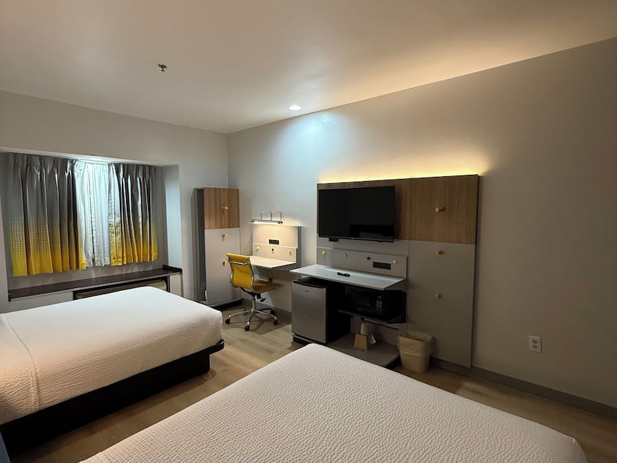 A modern hotel room with two beds, a desk, a chair, a television, and a window with curtains.