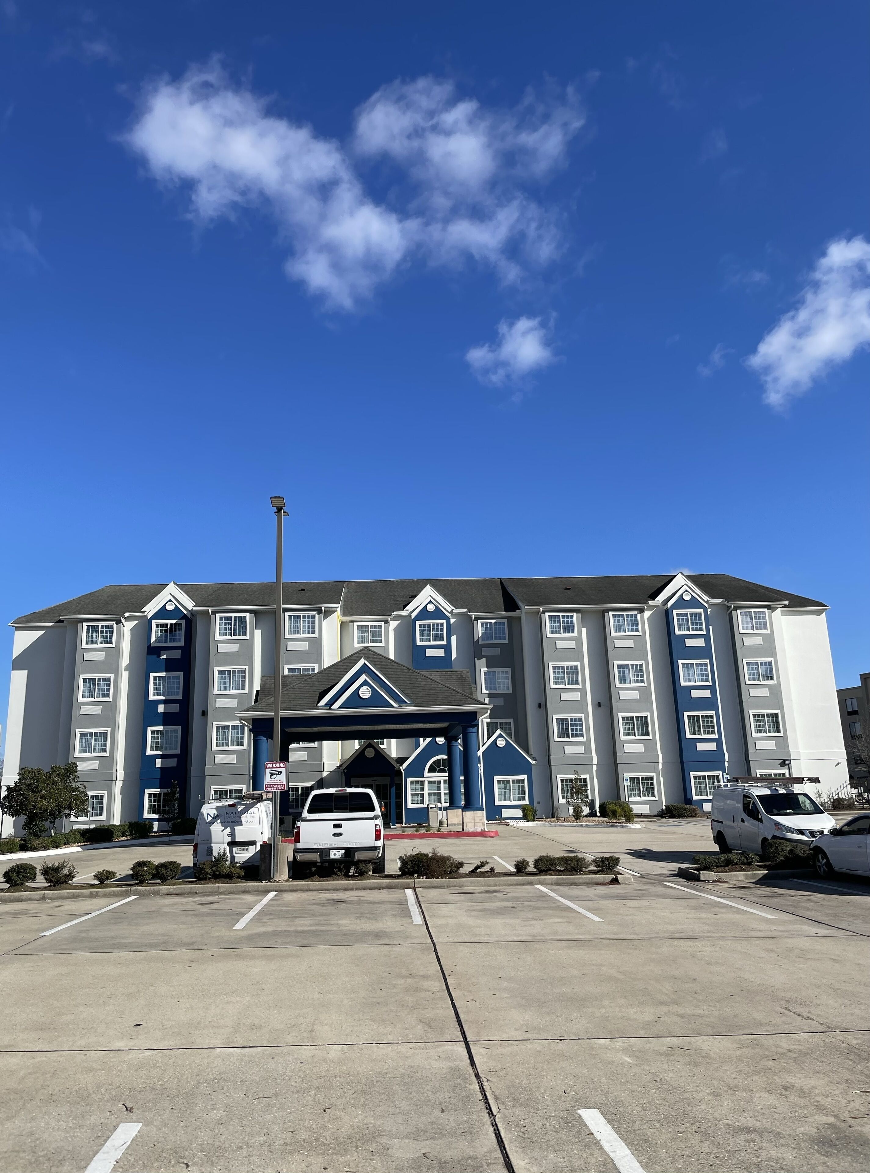 A multi-story hotel with a central entrance, parking lot, and a clear blue sky.