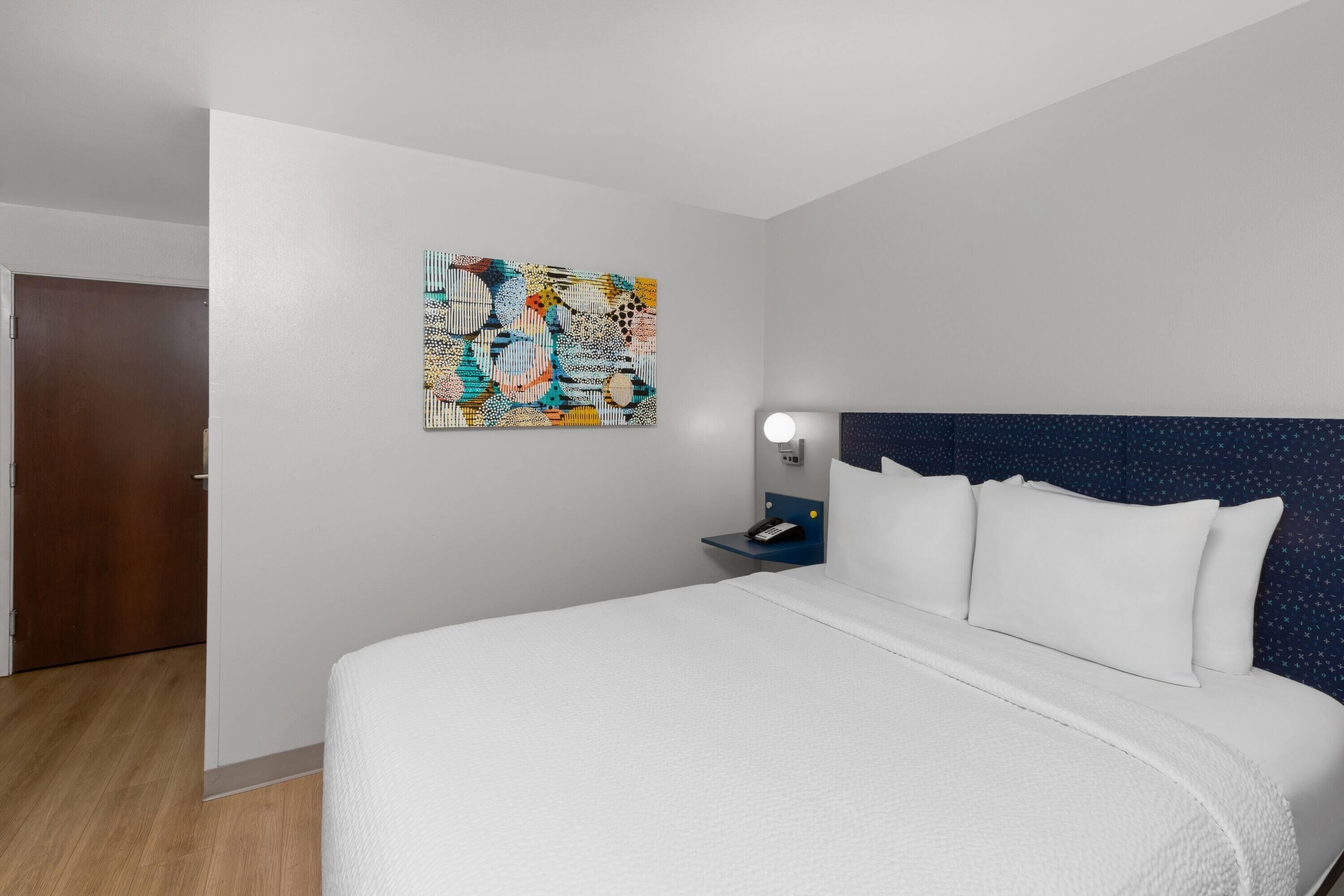 Quarto de hotel com uma cama grande, mesa de cabeceira com abajur, obra de arte na parede e uma porta.