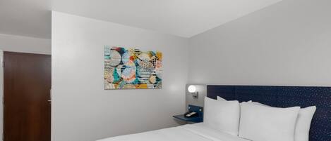 Quarto de hotel com uma cama grande, mesa de cabeceira com abajur, obra de arte na parede e uma porta.