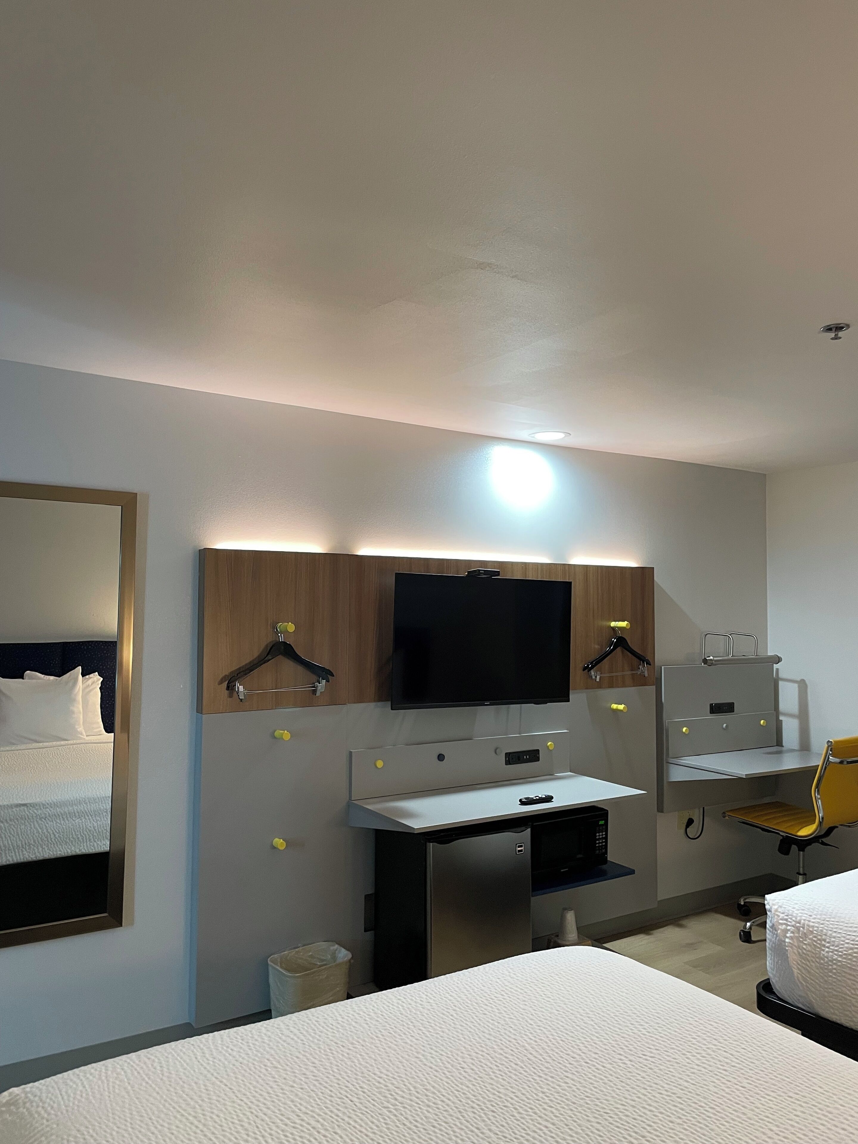 A hotel room with a bed, a desk with a computer, a television mounted above the desk, and a chair.