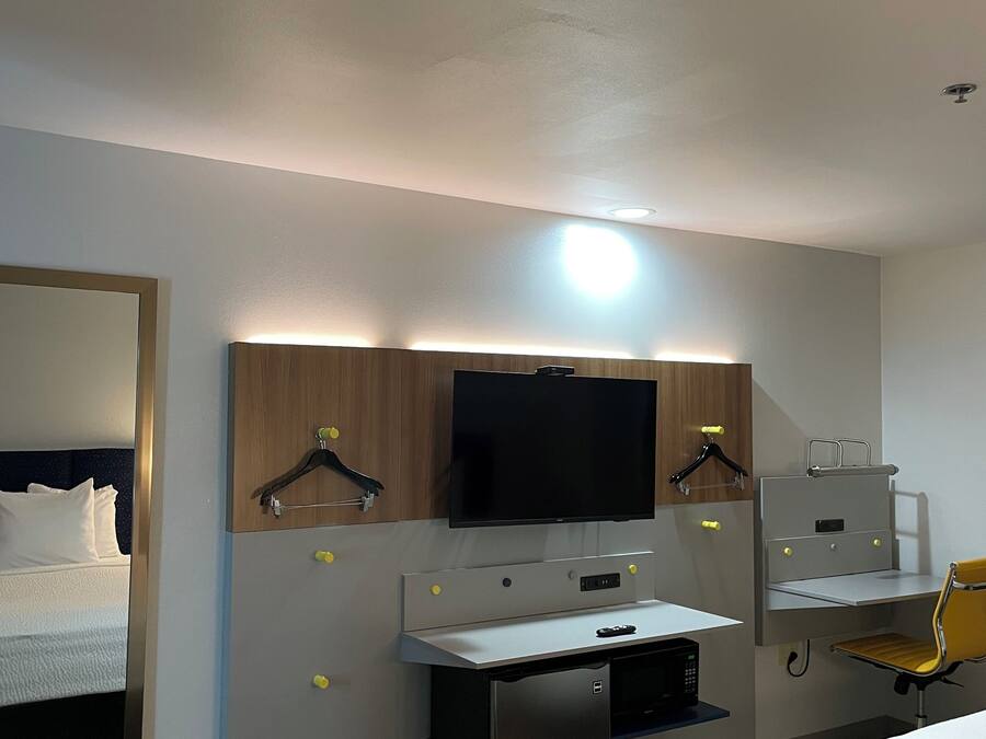 A hotel room with a bed, a desk with a computer, a television mounted above the desk, and a chair.