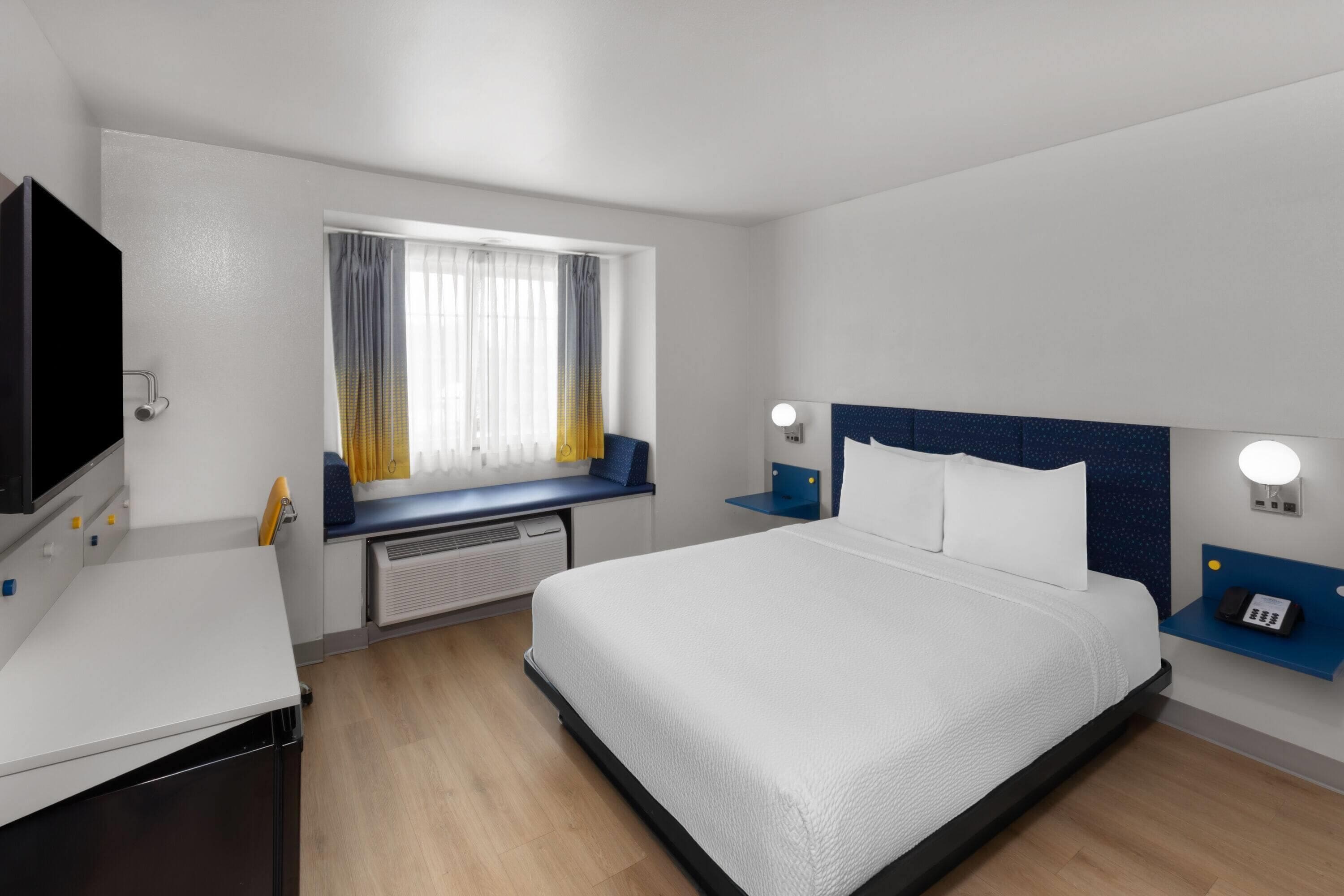 A hotel room with a bed, a desk, a TV, a window with curtains, and a nightstand with a phone.