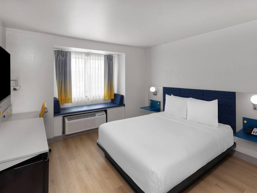 A hotel room with a bed, a desk, a TV, a window with curtains, and a nightstand with a phone.