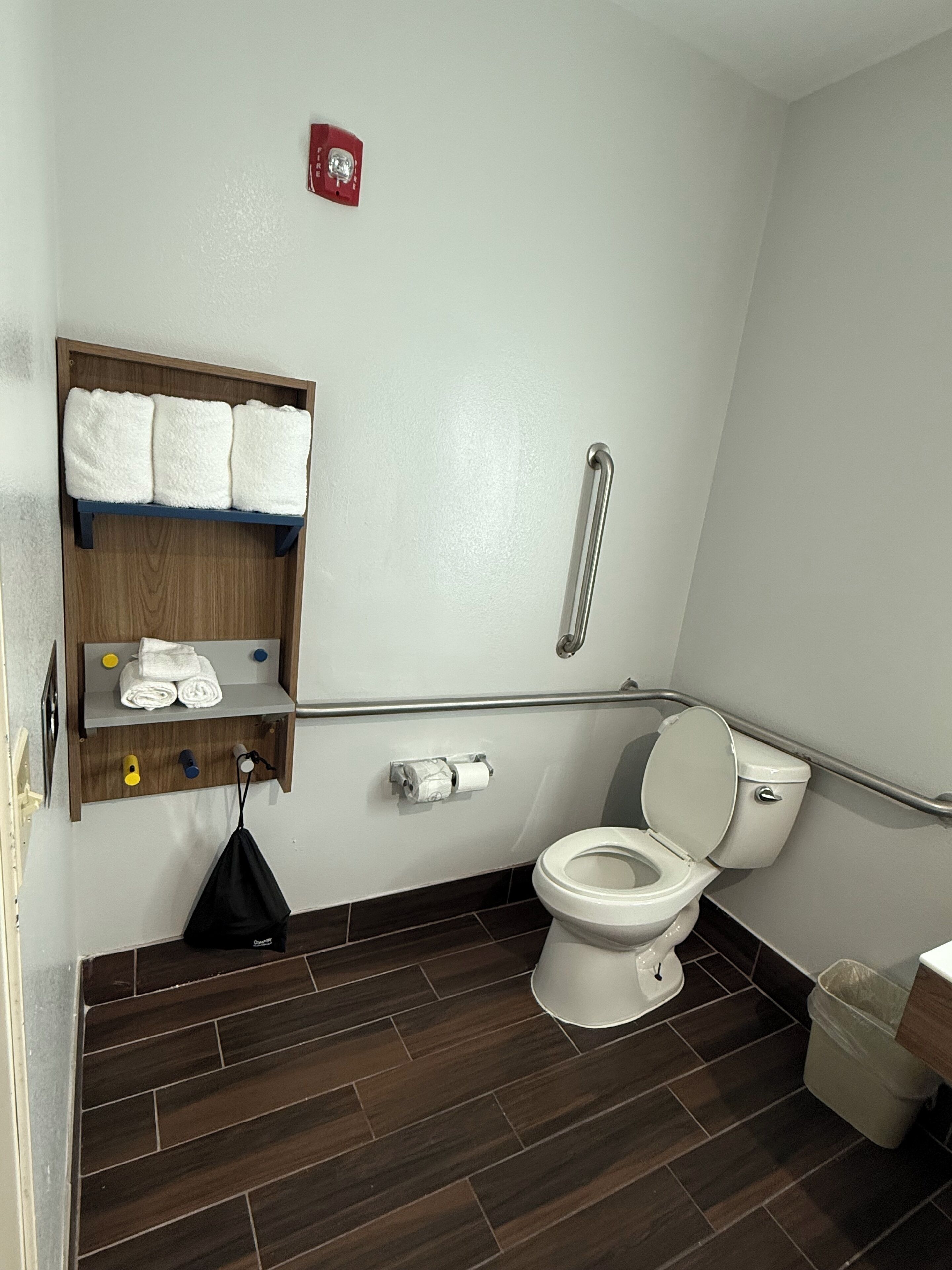 A bathroom with a toilet, a shelf with towels, a handrail, and a trash bin.