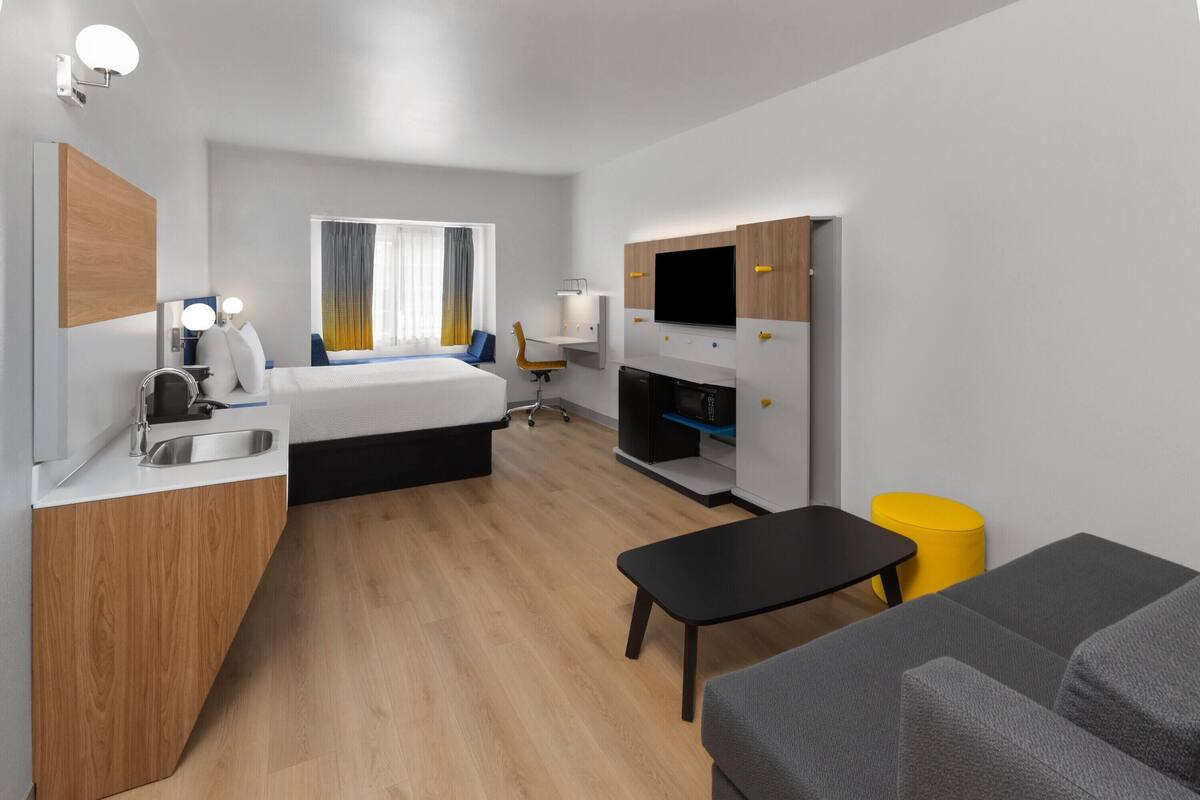 A modern hotel room with a bed, a desk, a TV, a sink, and a small dining area.