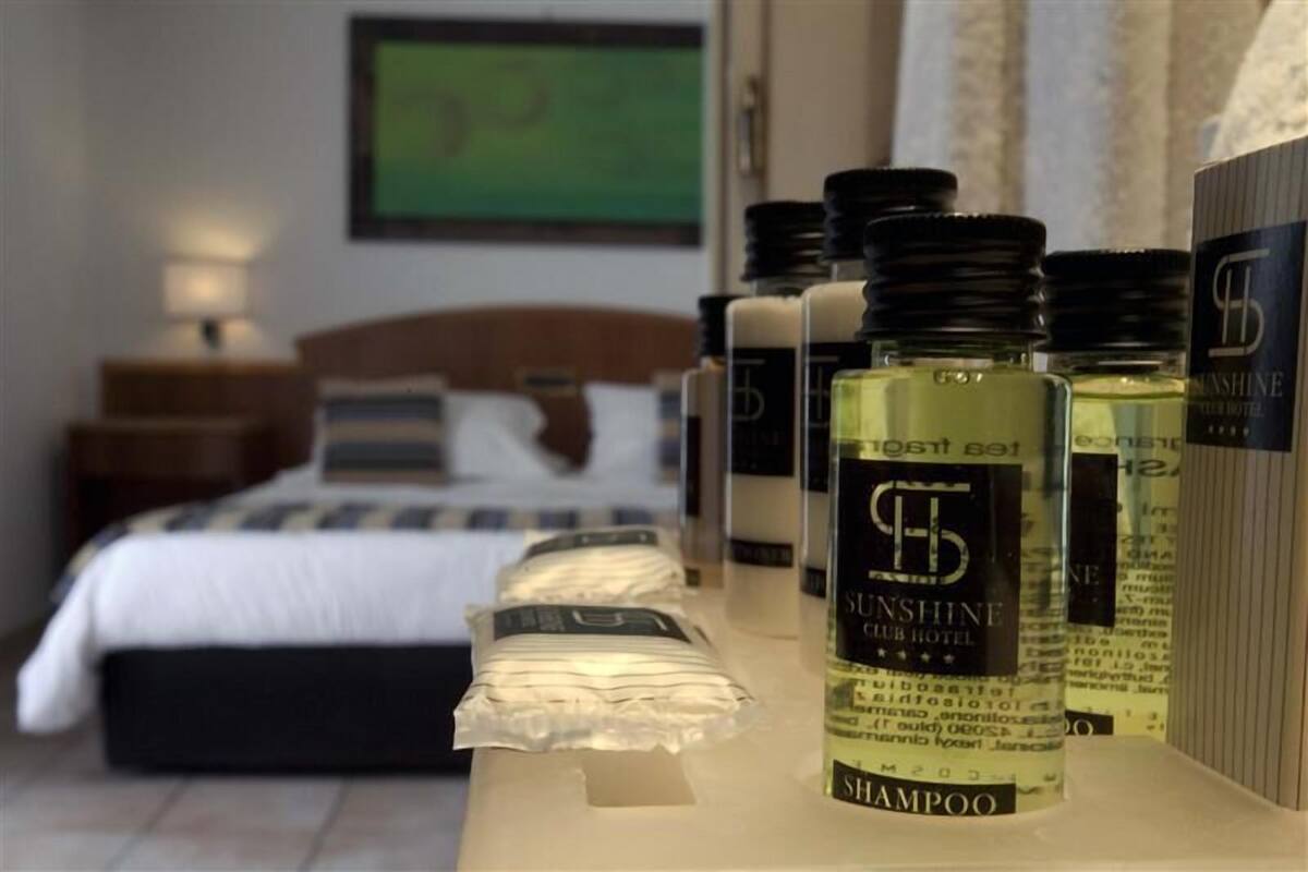 Et hotelværelse med en pænt redt seng, et natbord med en lampe, og flere flasker Sunshine Club Hotel shampoo på en hylde.