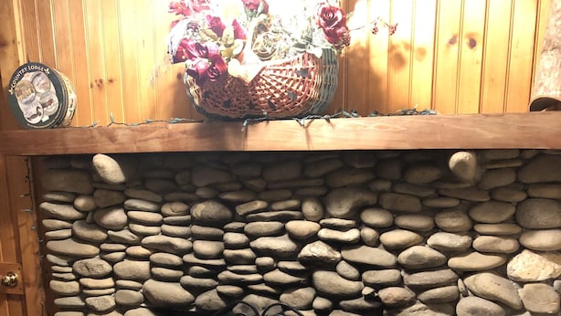 Ein Kamin aus Stein mit einem Metallsieb, eine Blumenkorb darauf und ein Holzstoß links daneben.
