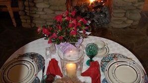 Ein Steinkamin mit Feuer, umgeben von einem Tisch, der mit Tellern, einer Kerze und einem Blumenstrauß gedeckt ist.