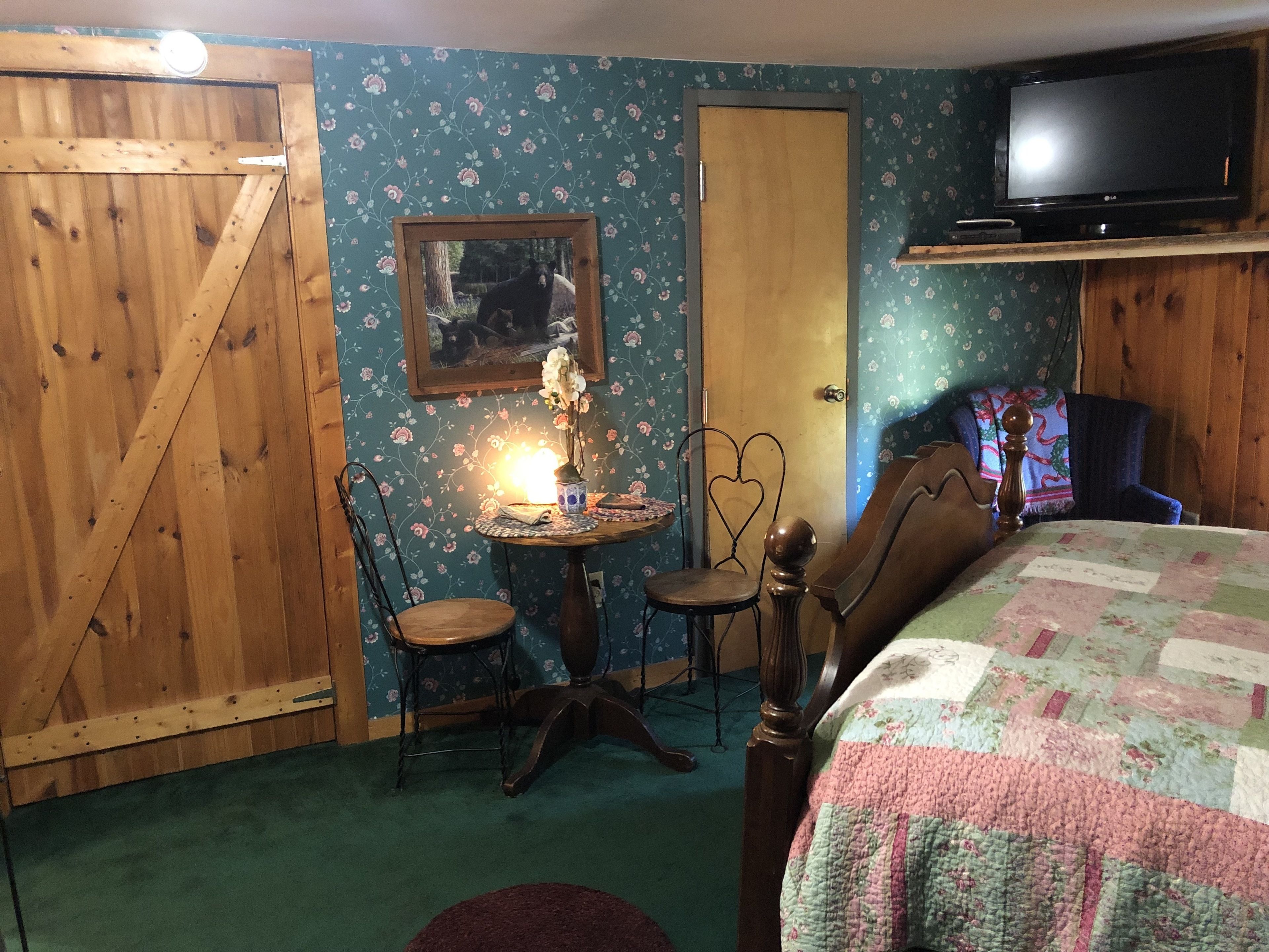 A cozy room with a wooden door, a floral wallpaper, a bed with a quilt, a television, and a small table with a lamp and a vase.
