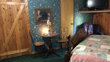 A cozy room with a wooden door, a floral wallpaper, a bed with a quilt, a television, and a small table with a lamp and a vase.