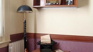 Une pièce d’angle avec un bureau en bois, une chaise rouge, une machine à écrire ancienne, un radiateur et une étagère contenant des bouteilles et un tableau encadré.