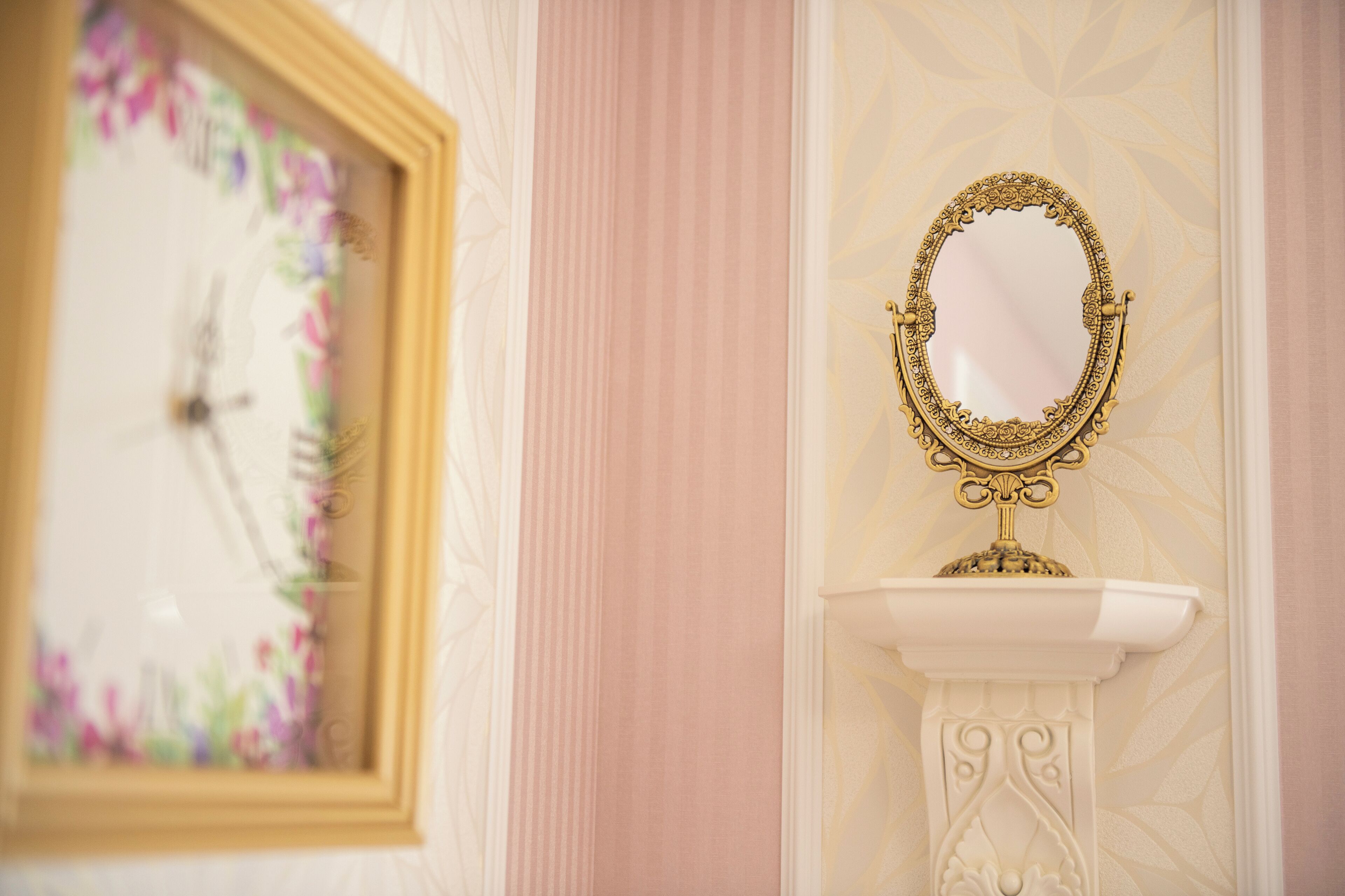 Un miroir encadré, posé sur un piédestal, est adossé à un mur orné d’un motif floral.