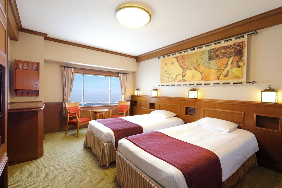 A hotel room with two beds, a desk, a chair, a window with curtains, a map on the wall, and a wooden cabinet.