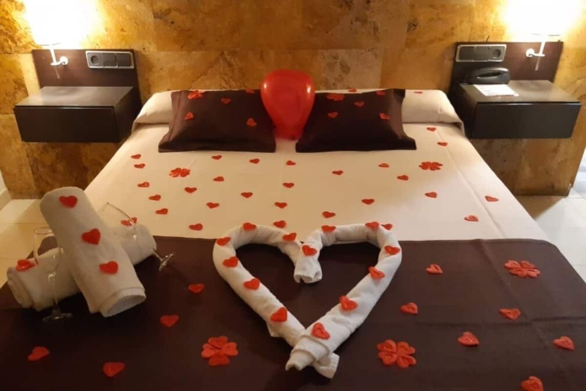 A hotel room with a bed, two bedside tables, a red balloon, heart-shaped decorations, and a heart-shaped towel arrangement.