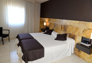 Habitación de hotel con dos camas, una silla, una mesita de noche con lámpara y una pared decorativa de piedra.