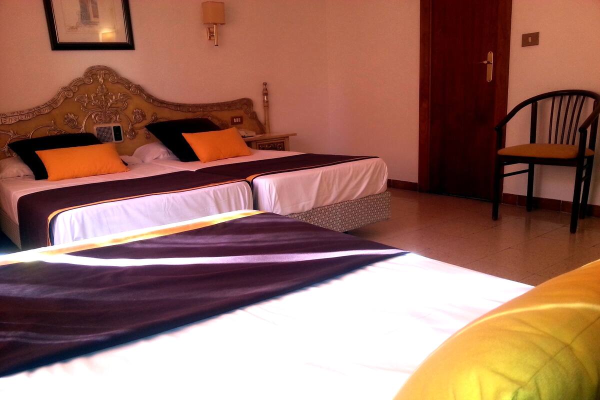 Habitación de hotel con dos camas, cabecera de madera, una silla y una mesita de noche con lámpara.