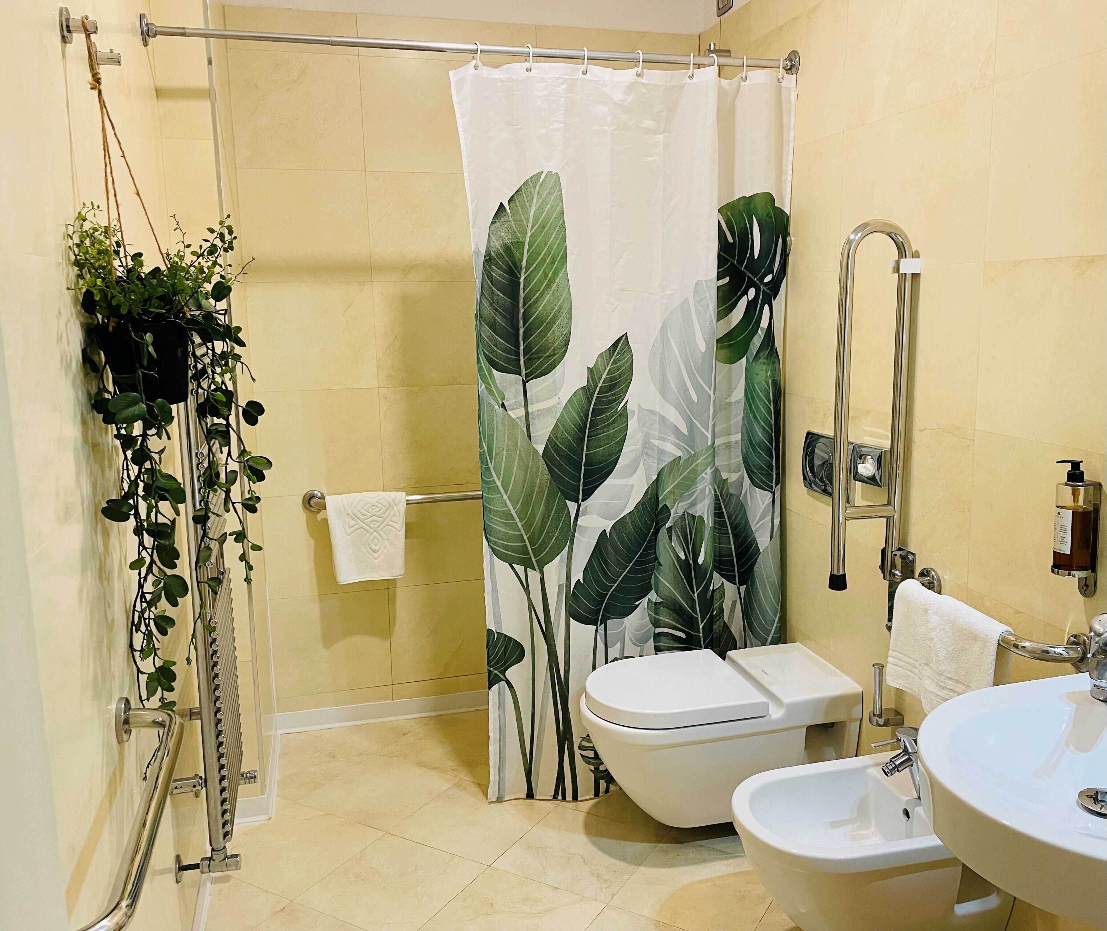 Casa de banho com cortina de chuveiro estampada com folhas verdes grandes, sanita, bidé, lavatório e toalheiro com toalha.