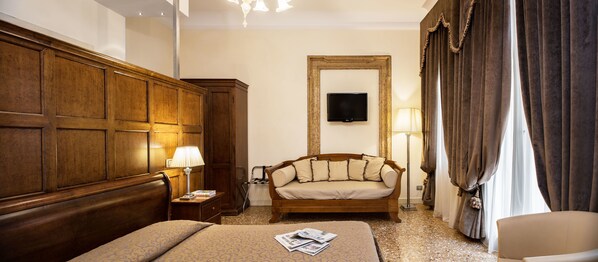 A hotel room with a wooden headboard, a bed with a patterned bedspread, a wooden nightstand with a lamp, a sofa, and a large mirror on the wall. - Hotel Dell'Opera (Venice)