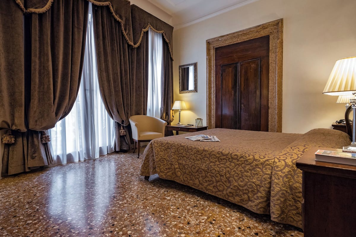 Une chambre d’hôtel avec un grand lit, une chaise, un bureau avec une lampe et une porte.