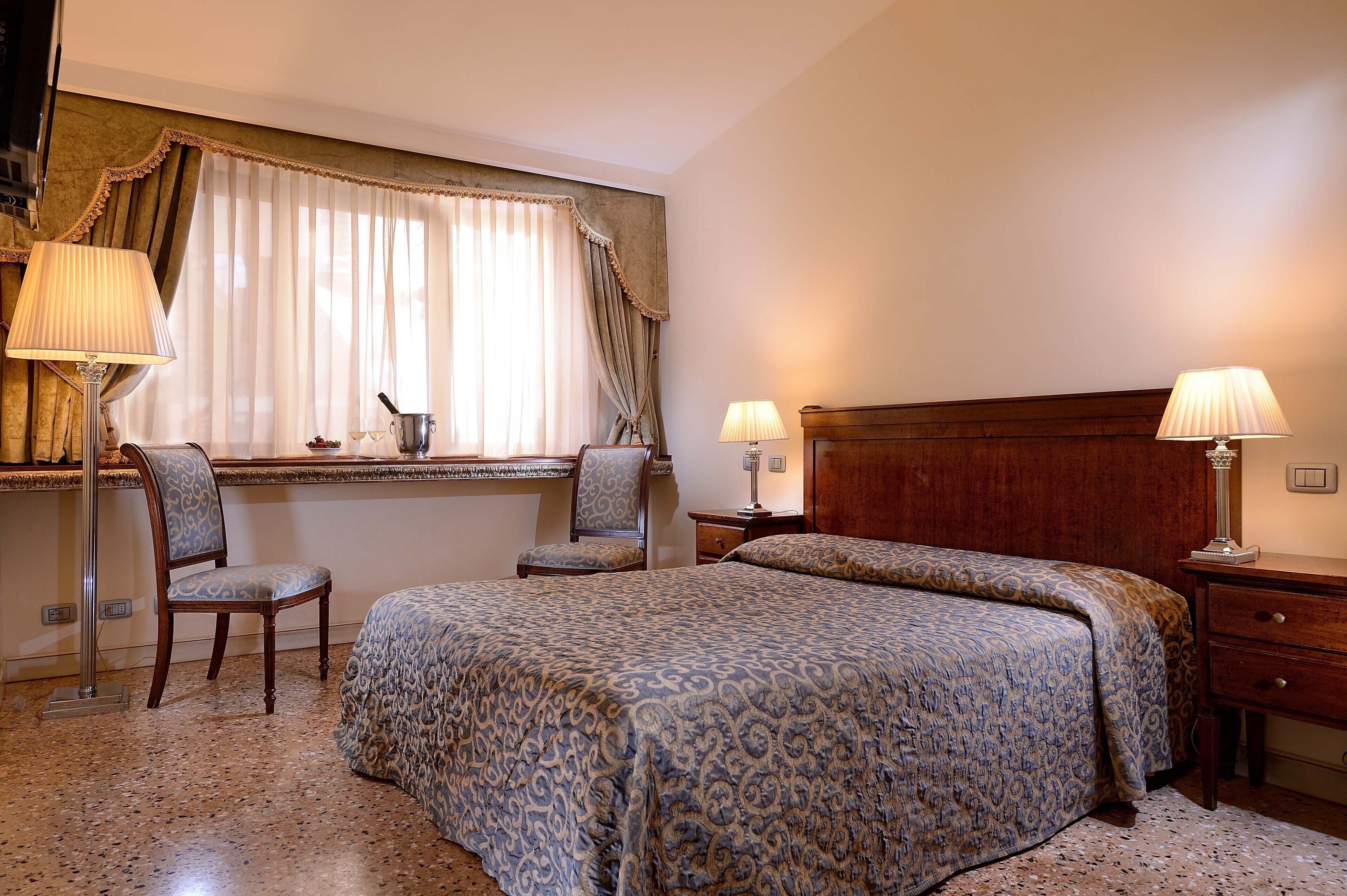 Une chambre d’hôtel avec un lit, deux chaises, un bureau, une lampe et une fenêtre avec des rideaux.