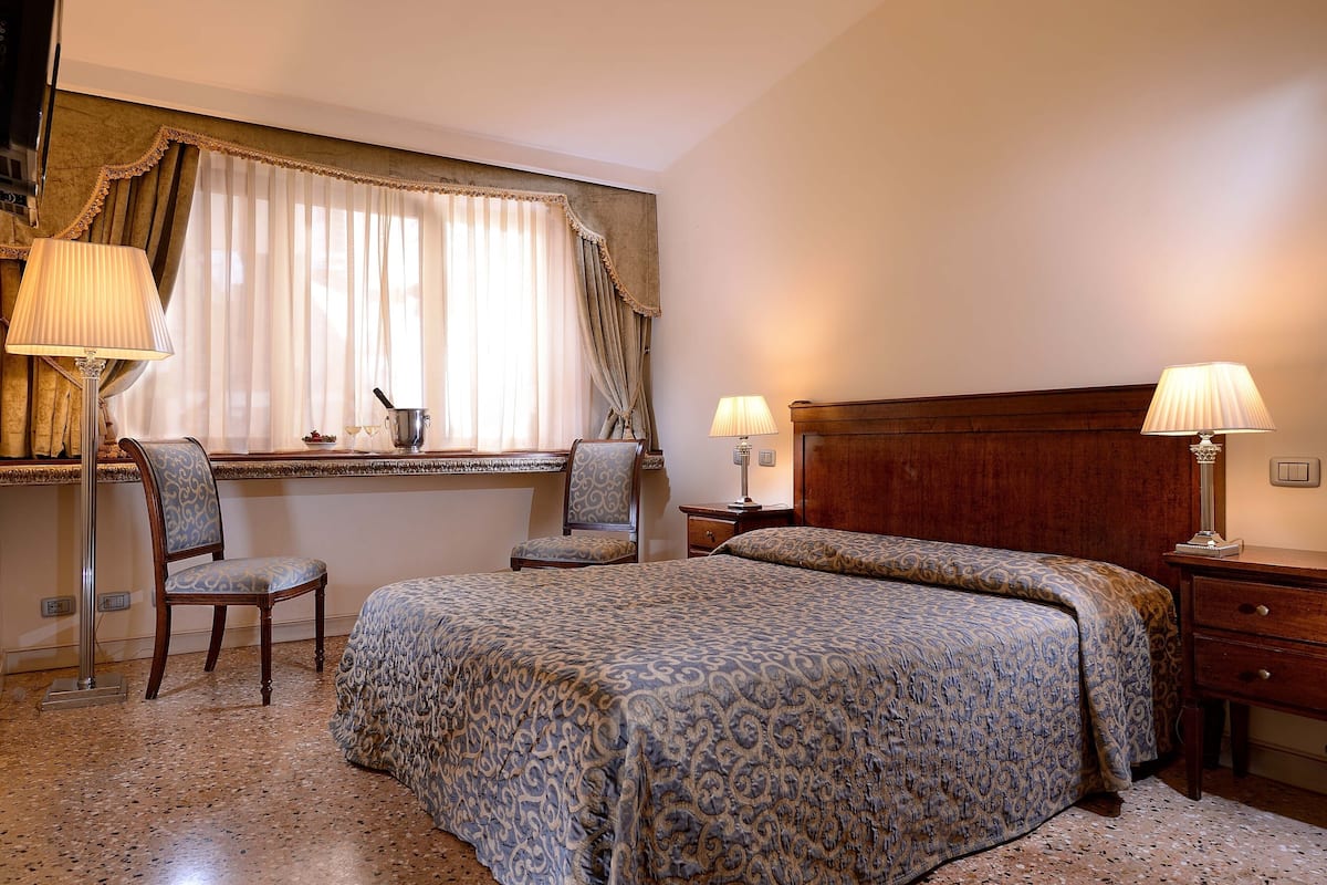 Une chambre d’hôtel avec un lit, deux chaises, un bureau, une lampe et une fenêtre avec des rideaux.