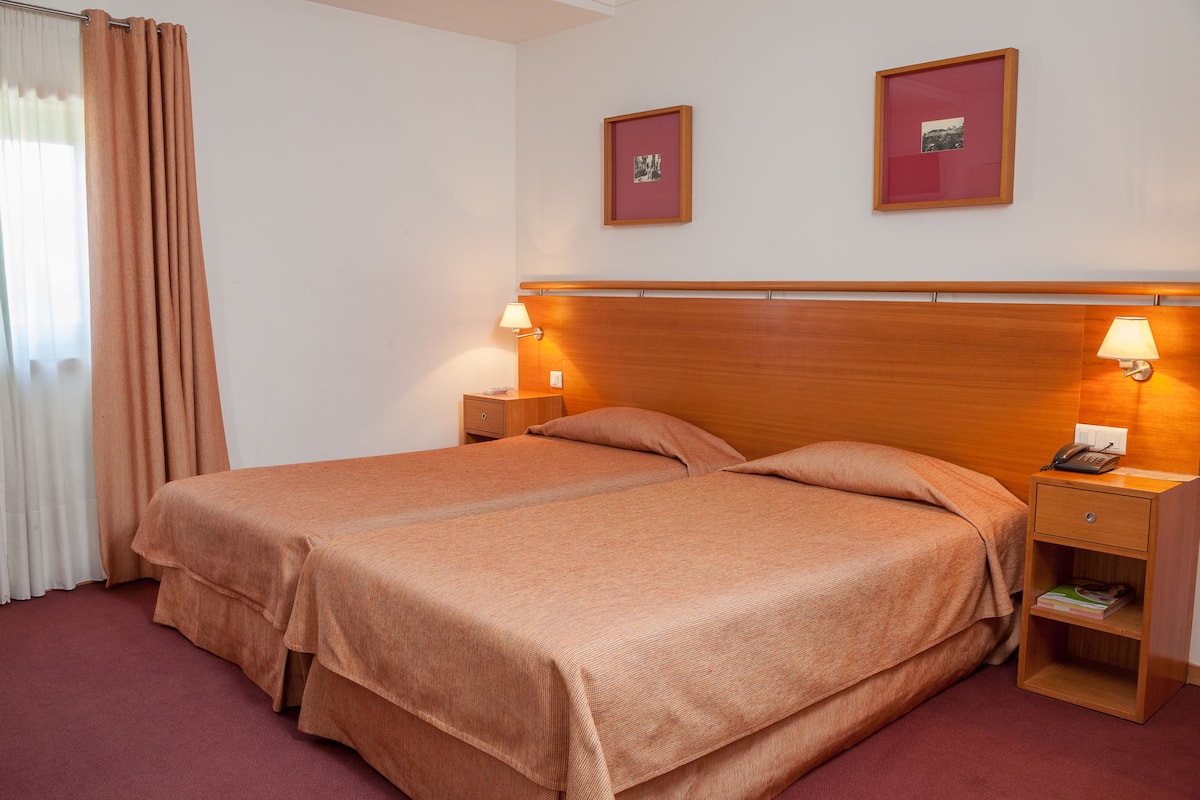 Una cama doble con cabecera de madera, dos mesitas de noche con lámparas, un cajón pequeño junto a la cama y dos cuadros enmarcados en la pared.