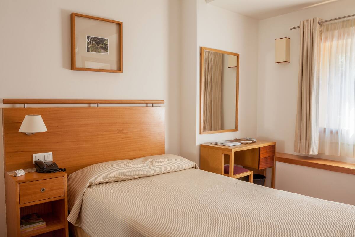 Habitación de hotel con cabecera de madera, mesita de noche con lámpara, escritorio pequeño y ventana con cortinas.