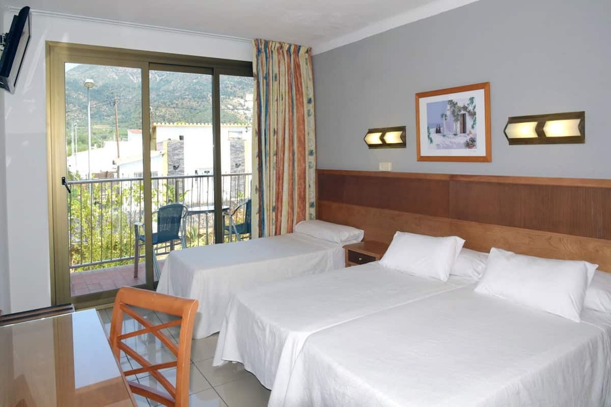 Habitación de hotel con dos camas, un balcón con vistas a edificios y montañas, y una mesa con sillas.