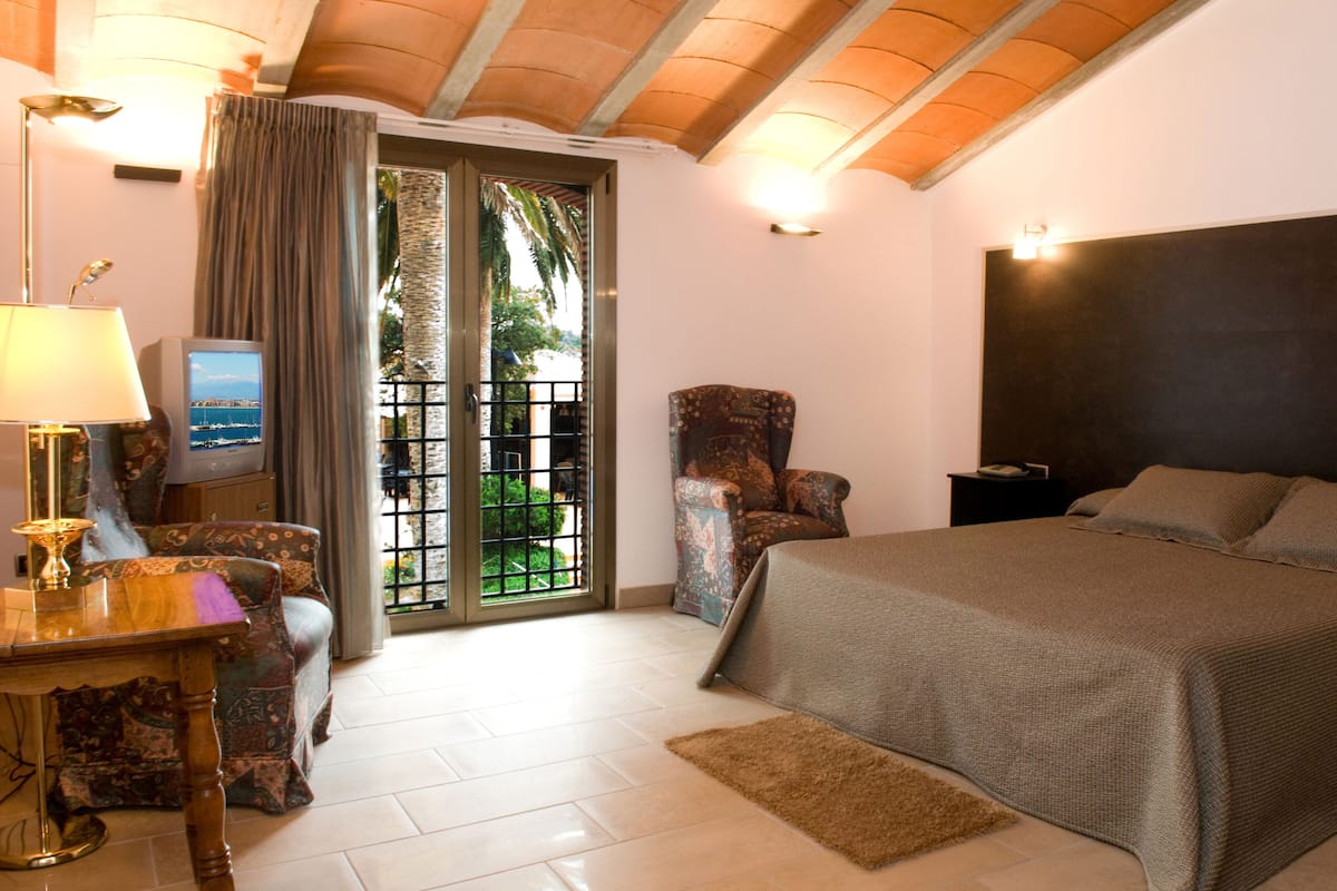 Habitación de hotel con cama, una silla, escritorio con lámpara, televisión y vista al exterior a través de una puerta.