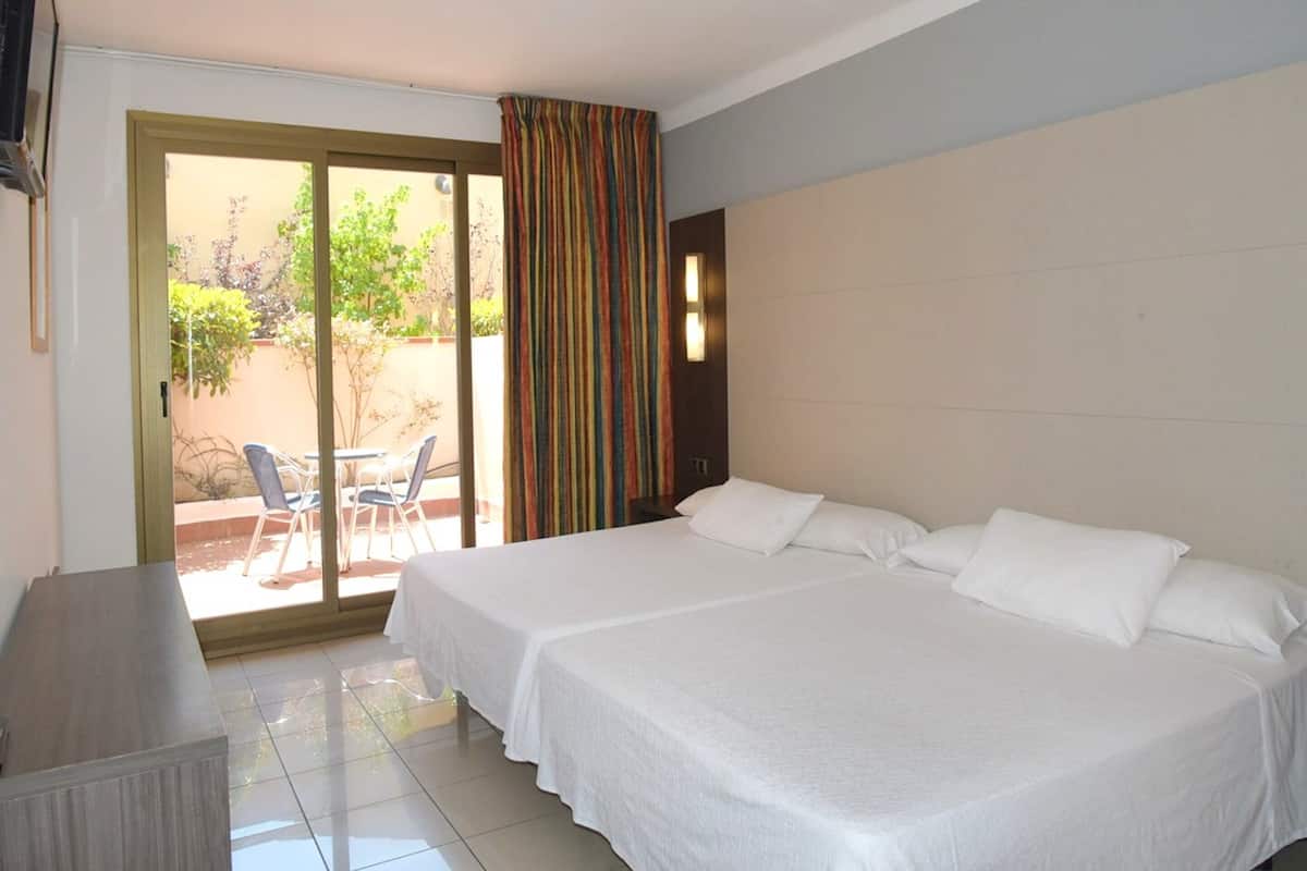 Habitación de hotel con dos camas, un escritorio y un ventanal corredero que da a un área exterior con sillas y una mesa.