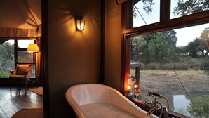 A room with a large bathtub, a chair, a lamp, and a view of the outdoors through a window. - Hamiltons Tented Camp (Bushbuckridge)