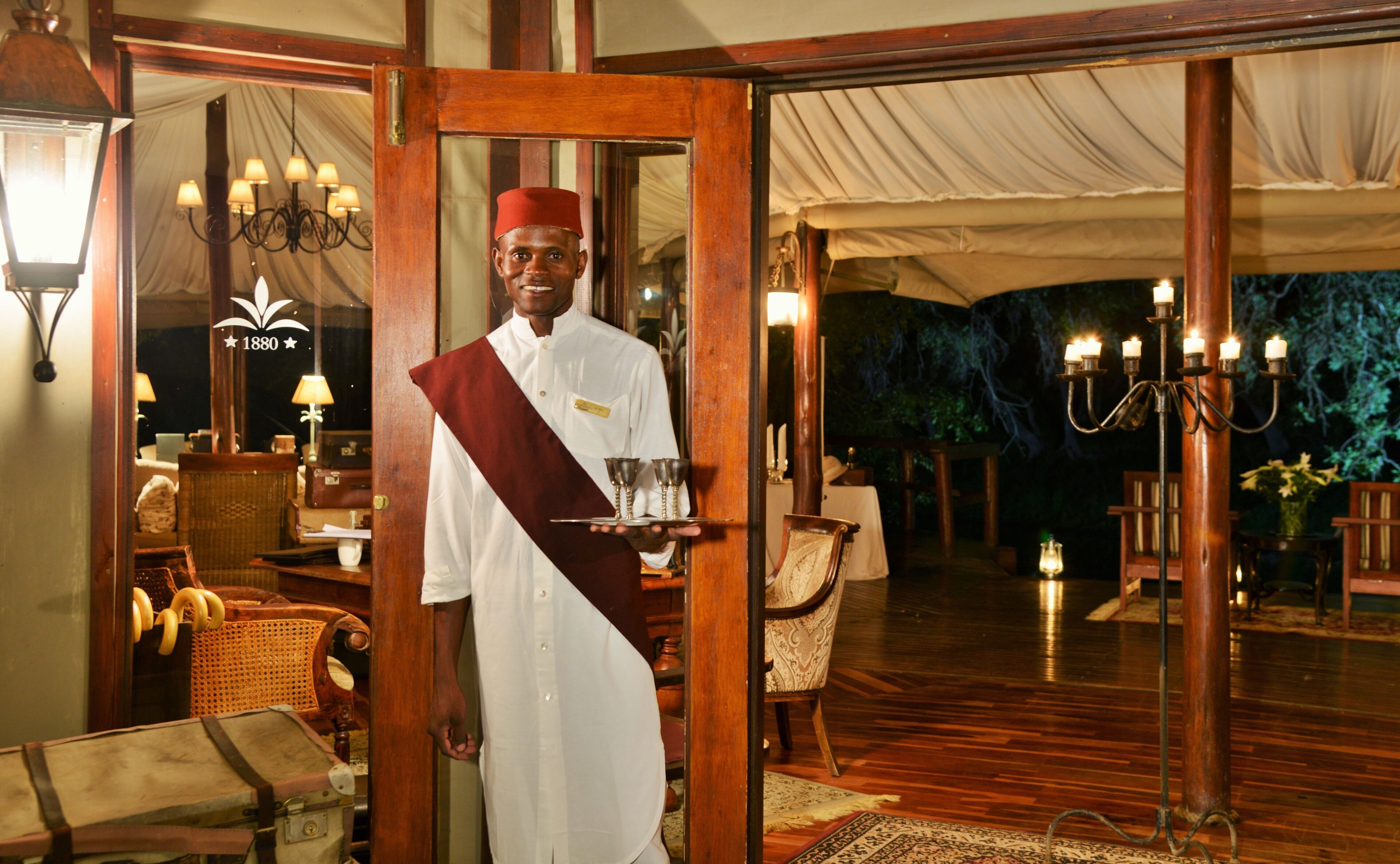 A waiter in traditional attire holding a tray with glasses, standing at a doorway with a wooden frame, a chandelier, and a tented area visible in the background.