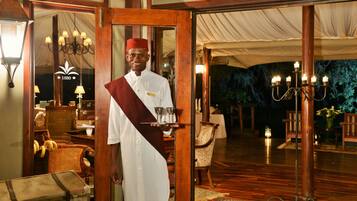 A waiter in traditional attire holding a tray with glasses, standing at a doorway with a wooden frame, a chandelier, and a tented area visible in the background.