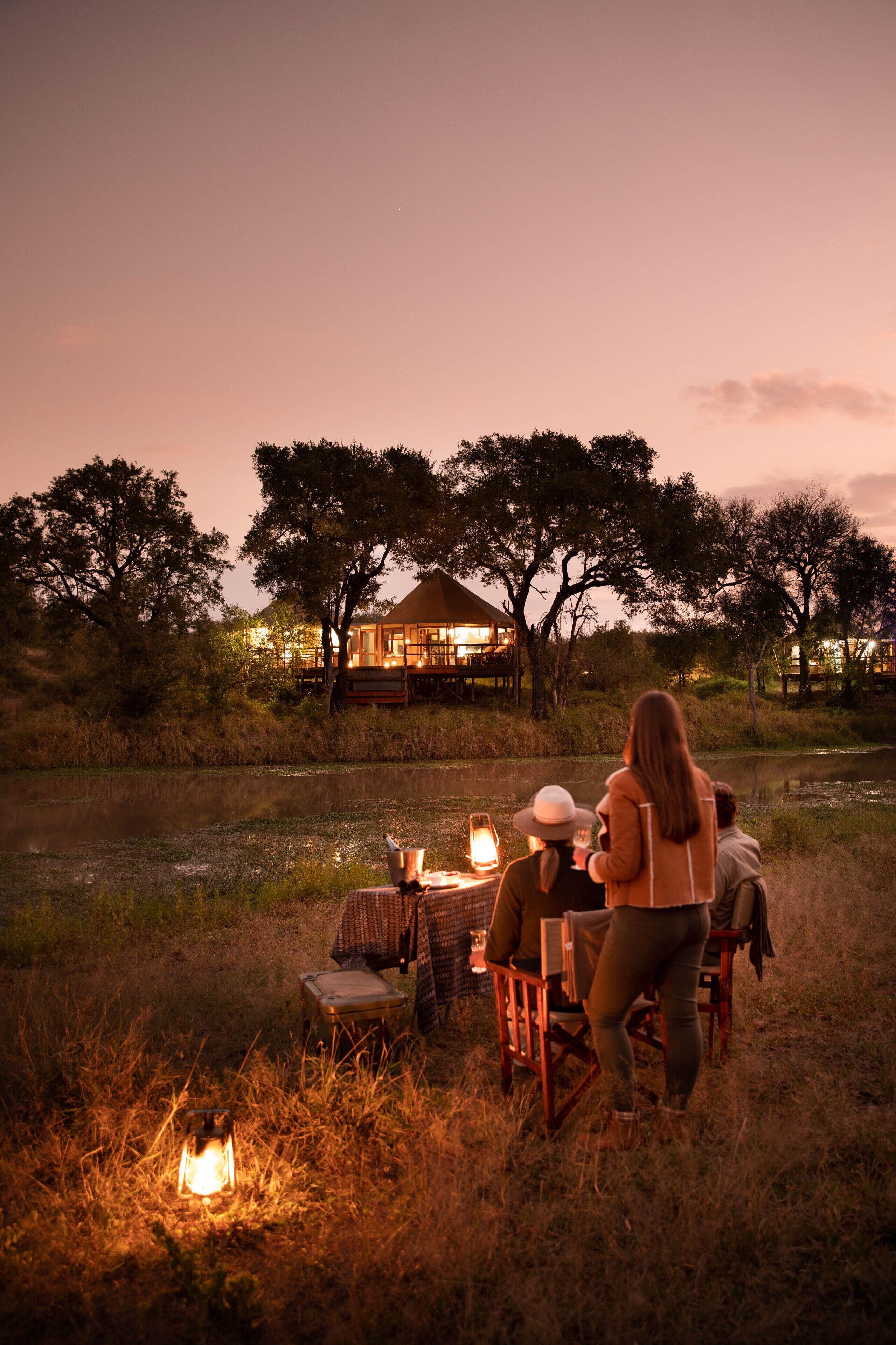 A group of people seated at a table outdoors with a lit lantern in the foreground, surrounded by tall grass and trees, with a building in the background.