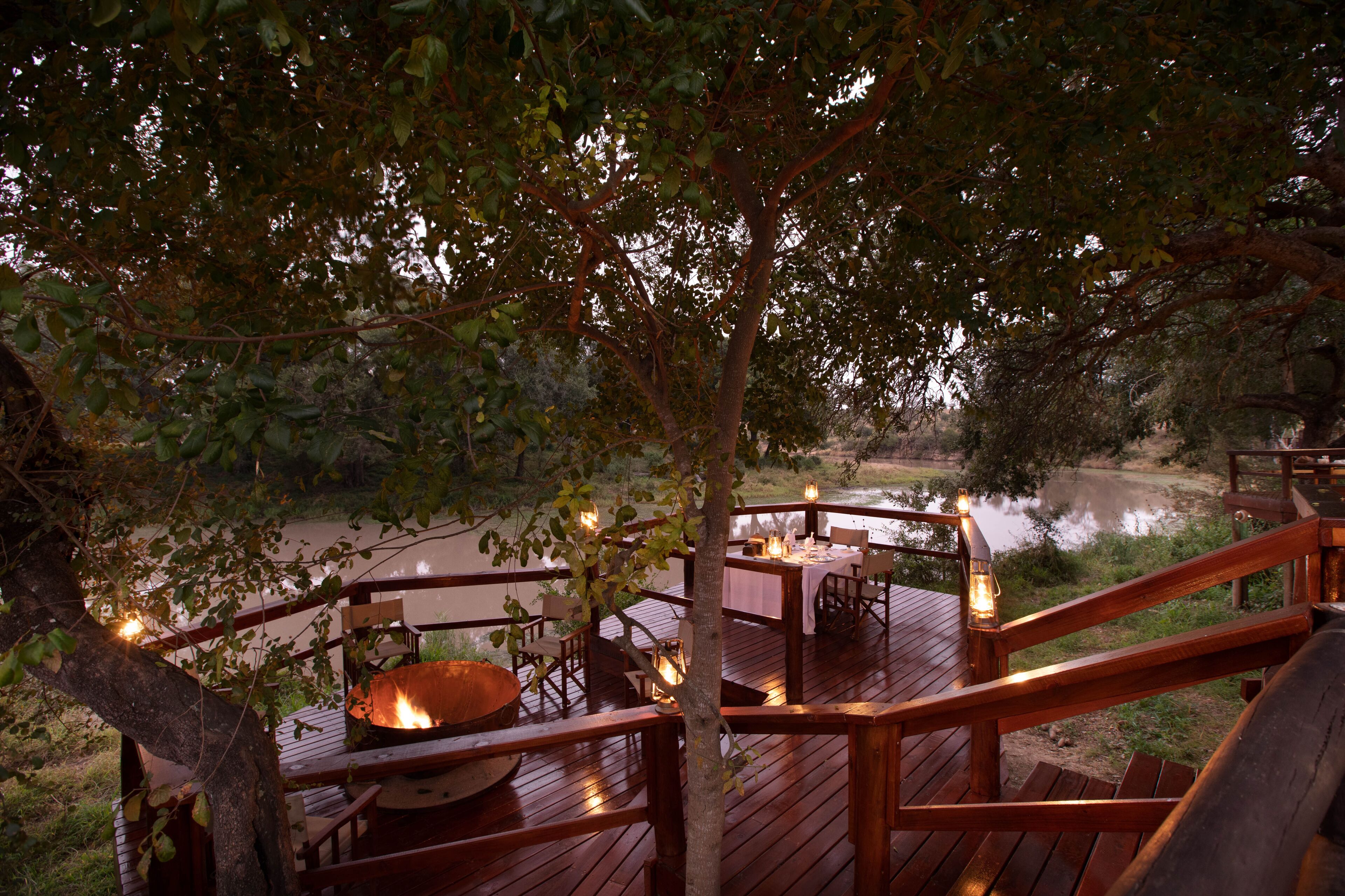 A wooden deck with tables and chairs, a fire pit, and a view of a river.