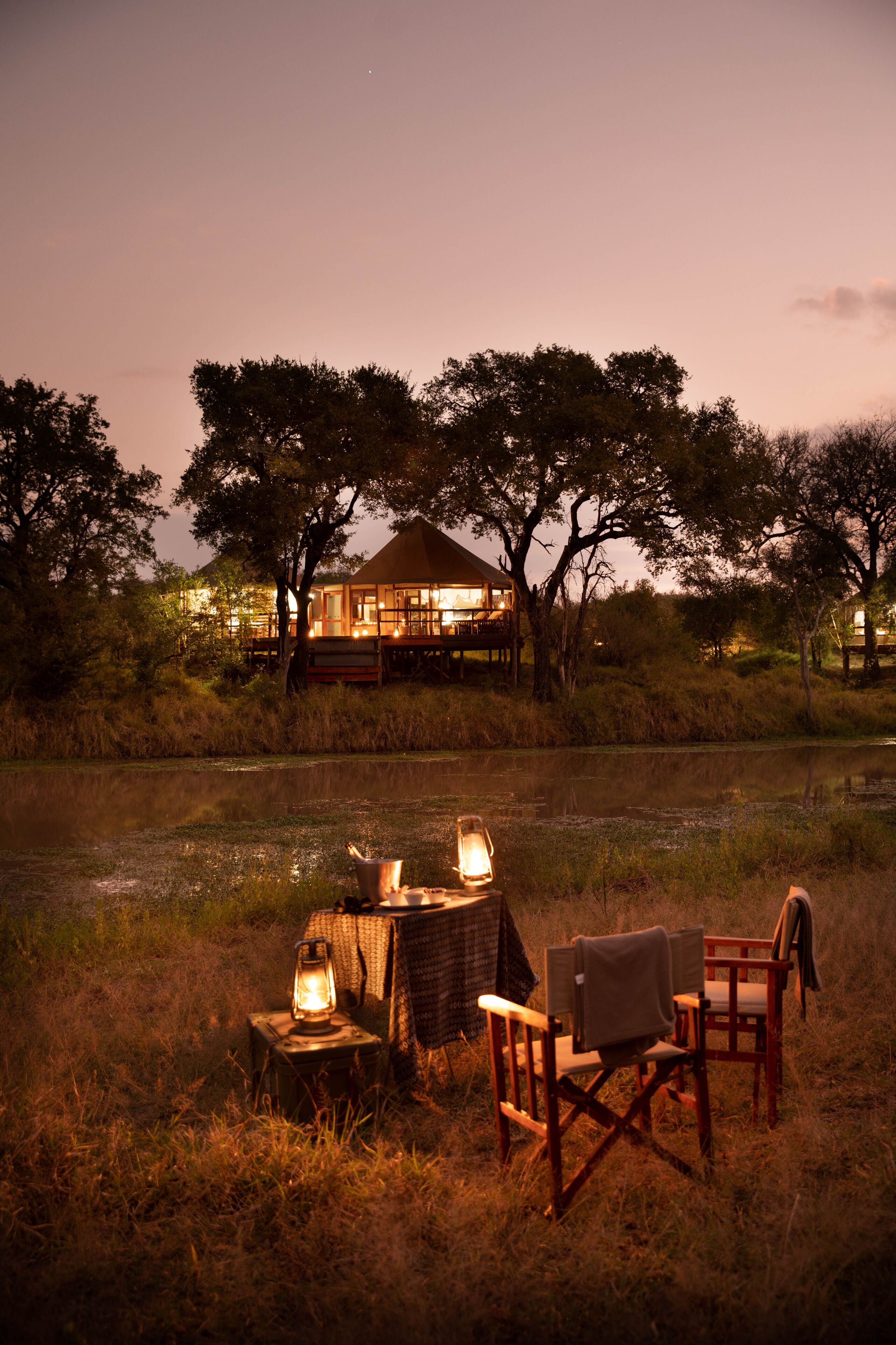 A rustic outdoor seating area with a lantern, a table, and two chairs, overlooking a body of water and a lodge in the background.
