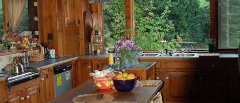 A rustic kitchen with wooden cabinets, a large wooden table, and a view of the surrounding greenery.
