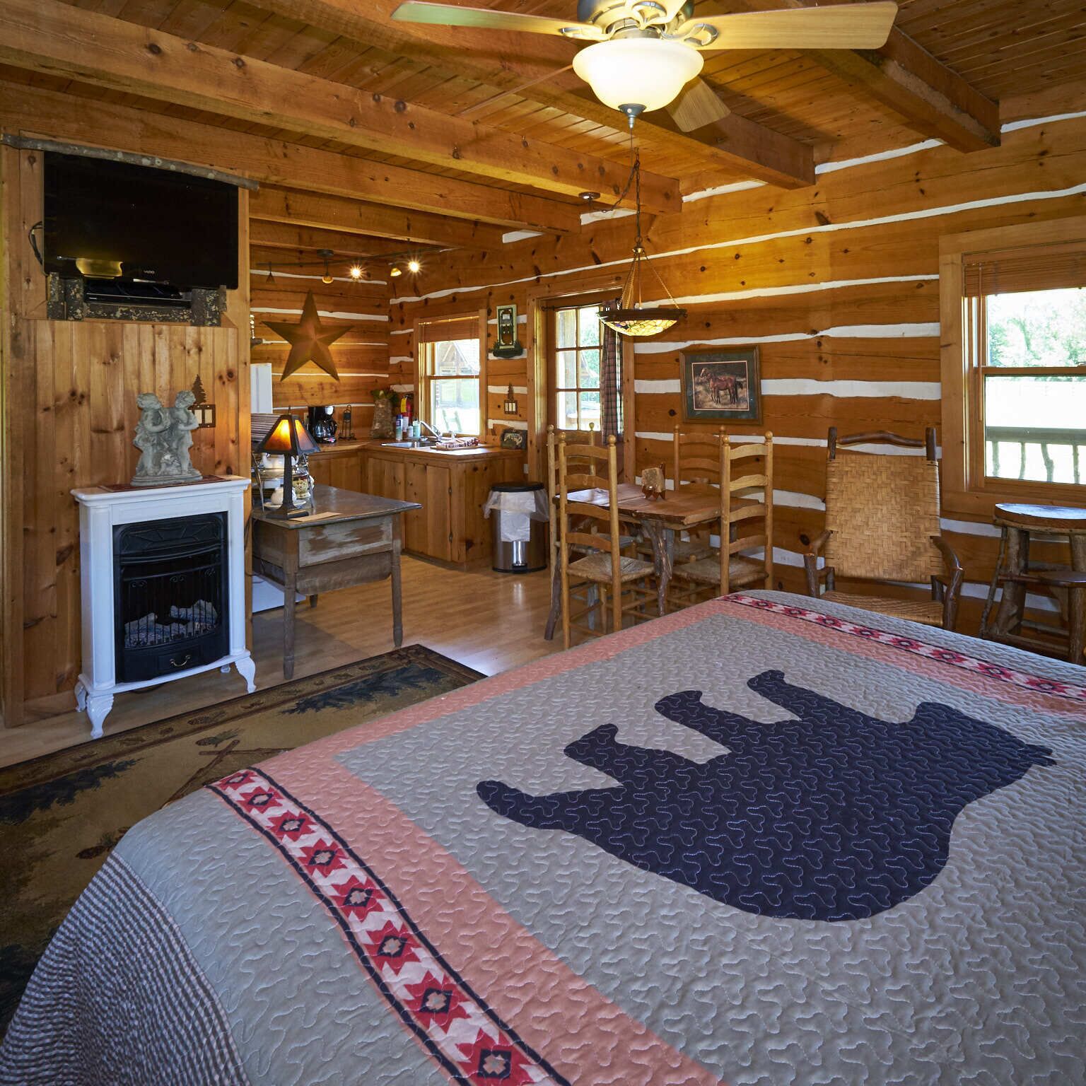 A log cabin interior with a bed, wooden walls, a fireplace, a dining area, and a kitchen.