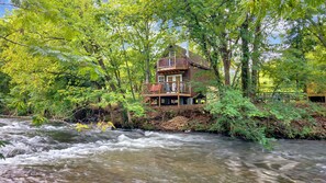 A wooden cabin with a deck, surrounded by dense forest and a flowing river. - Creekwalk Inn and Cabins (Cosby)