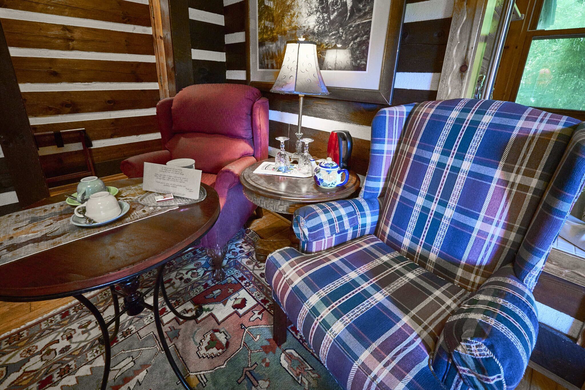 A cozy room with a plaid armchair, a red armchair, a small round table with a lamp, a tea set, and a rug.
