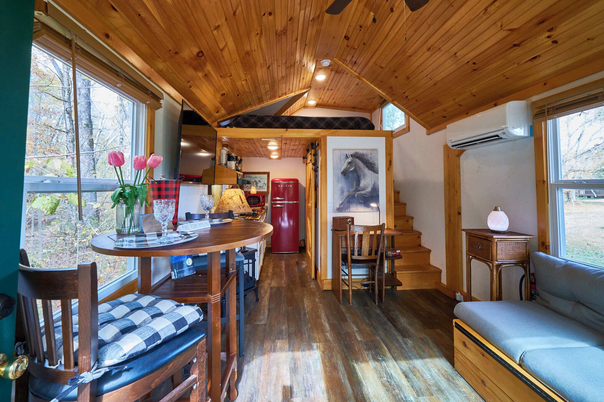 Interior acogedor de una cabaña con techo de madera, una zona de comedor con mesa y sillas, un sofá y una escalera que sube a un altillo.