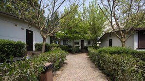A pathway leading to a row of white buildings with black doors, surrounded by greenery and trees. - Happy Village & Camping (Rome)