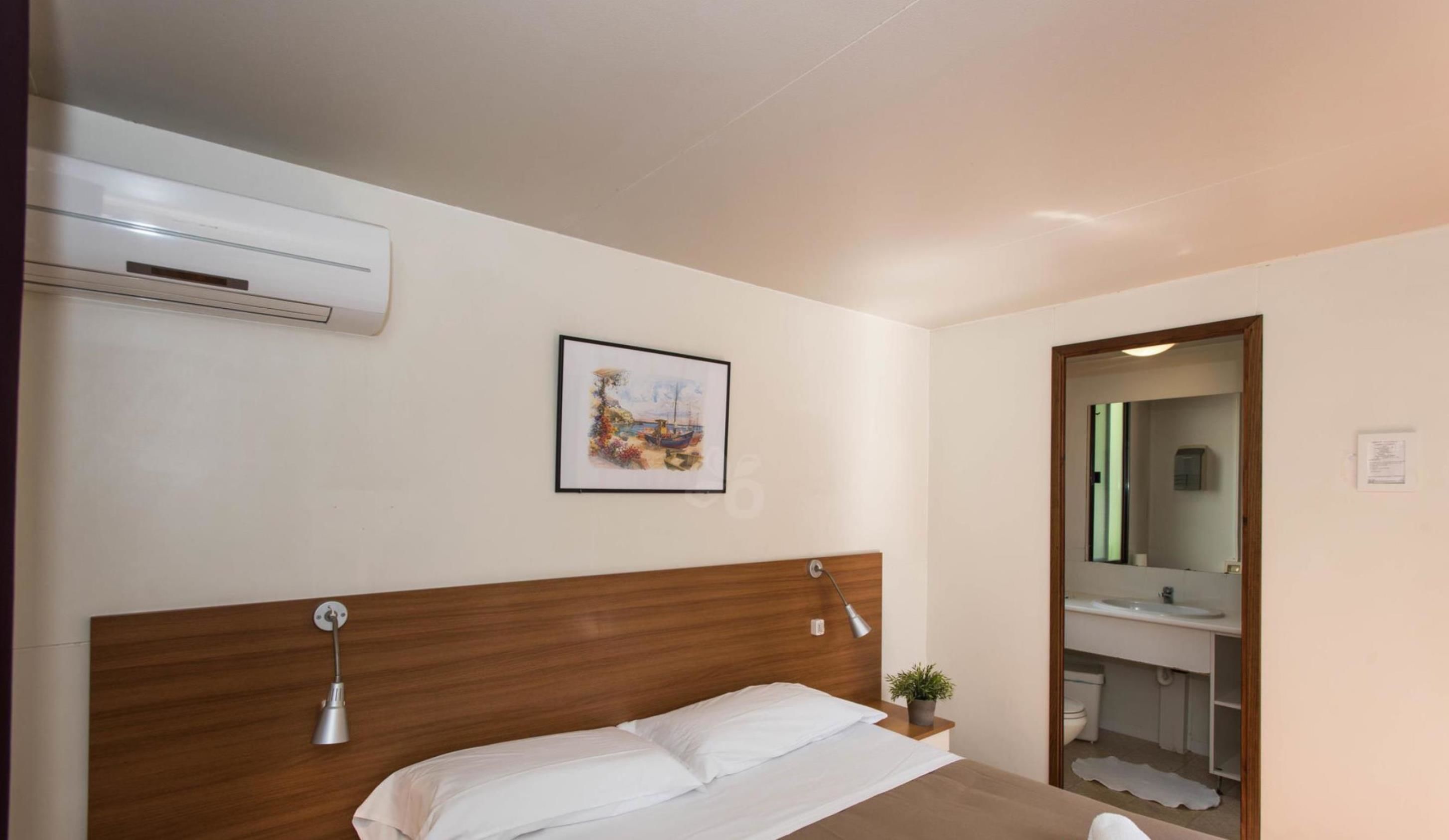 Ein Hotelzimmer mit einem hölzernen Kopfteil, einem Einzelbett, einer Nachttischlampe, einer Klimaanlage, einem gerahmten Gemälde und einem durch eine geöffnete Tür sichtbaren Badezimmer.
