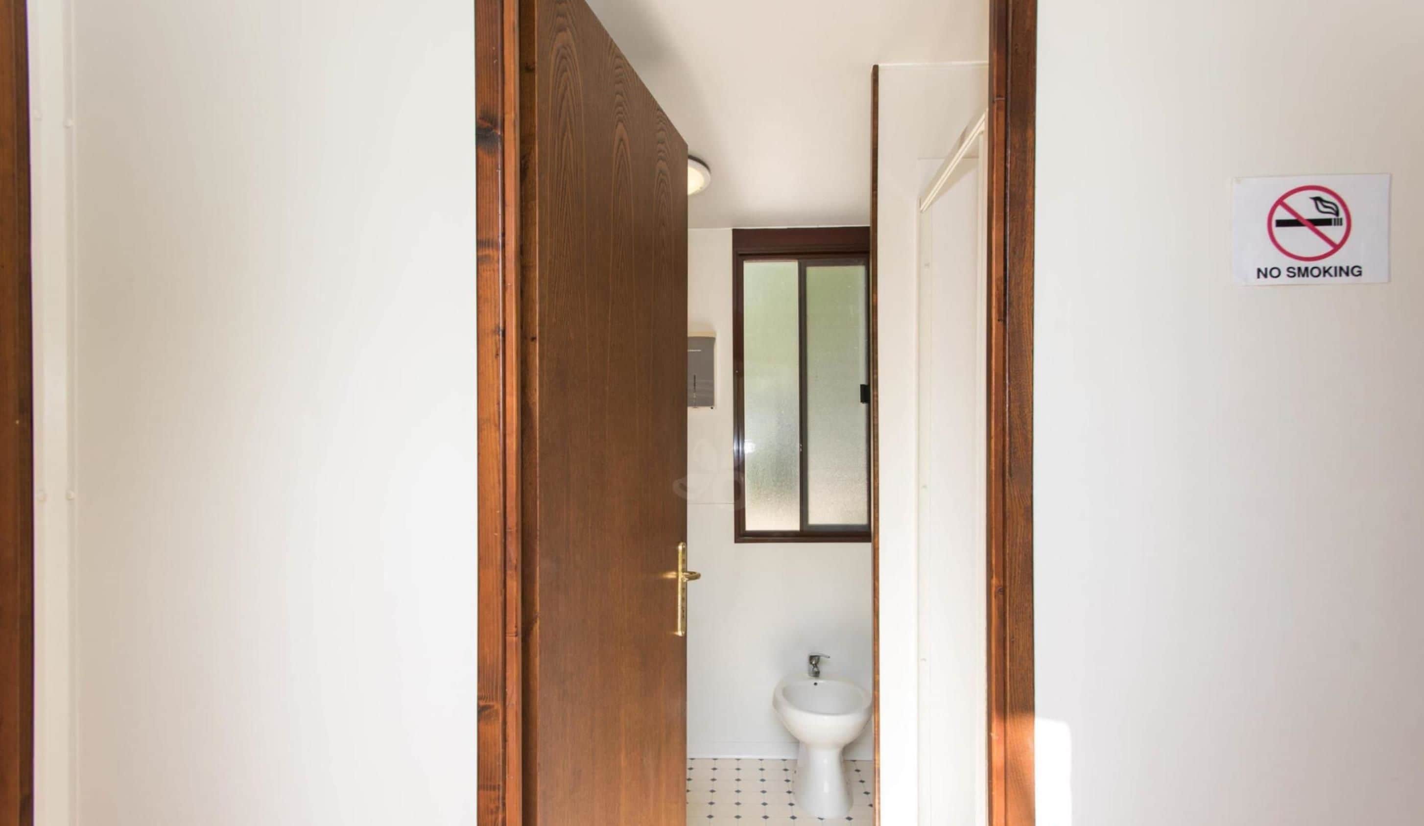 A narrow hallway leading to a bathroom with a toilet and bidet, a 'No Smoking' sign on the wall, and a wooden door.