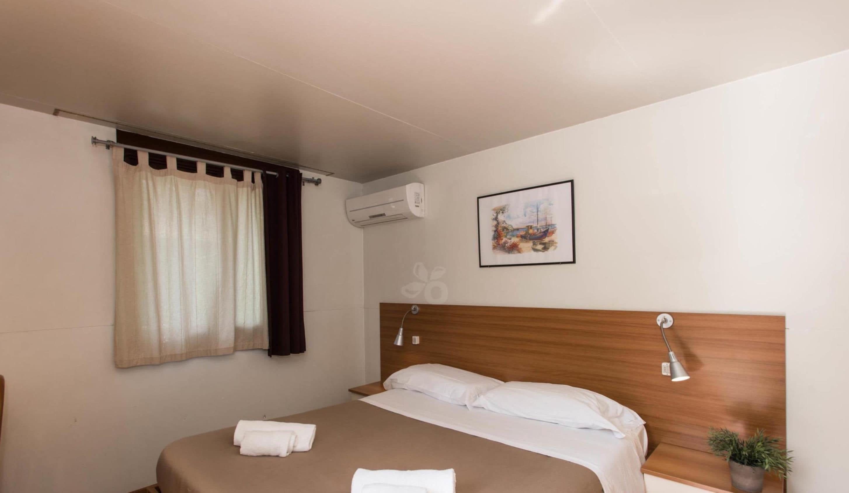 Une chambre d’hôtel avec un lit, une fenêtre avec des rideaux, un appareil de climatisation, une lampe fixée au mur, un tableau encadré et une petite plante en pot.