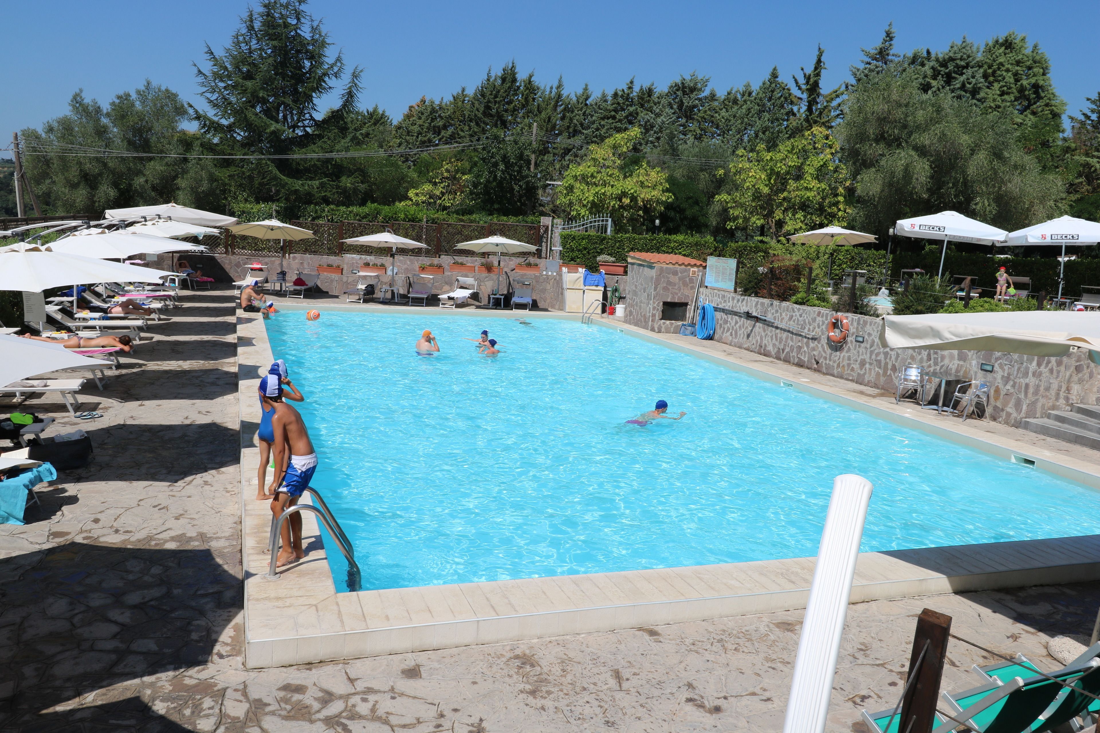 A swimming pool with multiple people swimming and lounging around. There are umbrellas, lounge chairs, and a lifebuoy visible.