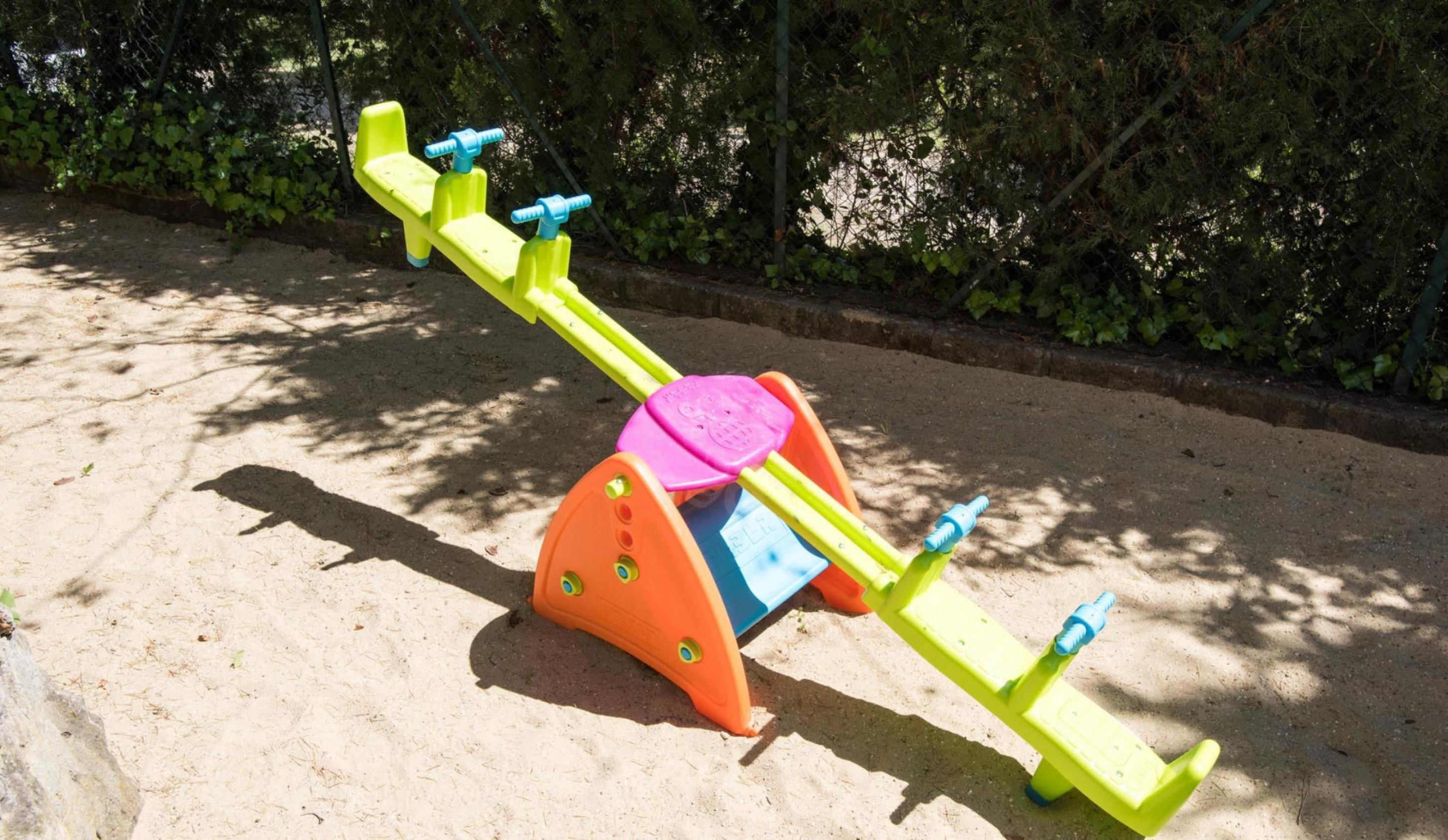 A seesaw with a pink seat and a blue base, set on sandy ground with trees in the background.